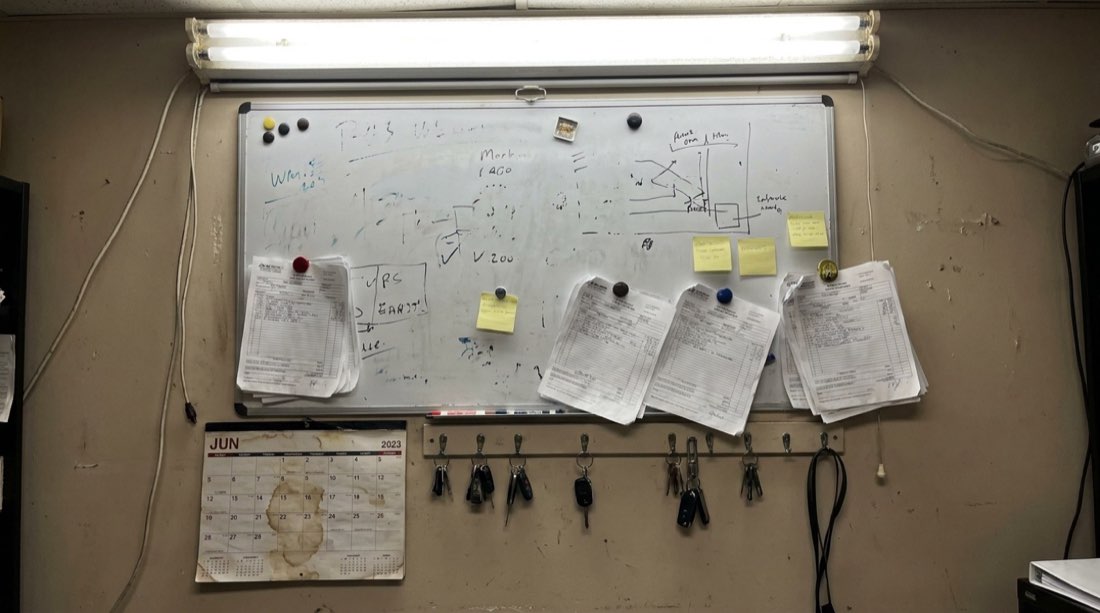 A real small-shop dispatch wall: chaotic whiteboard, taped work orders, keys on hooks.